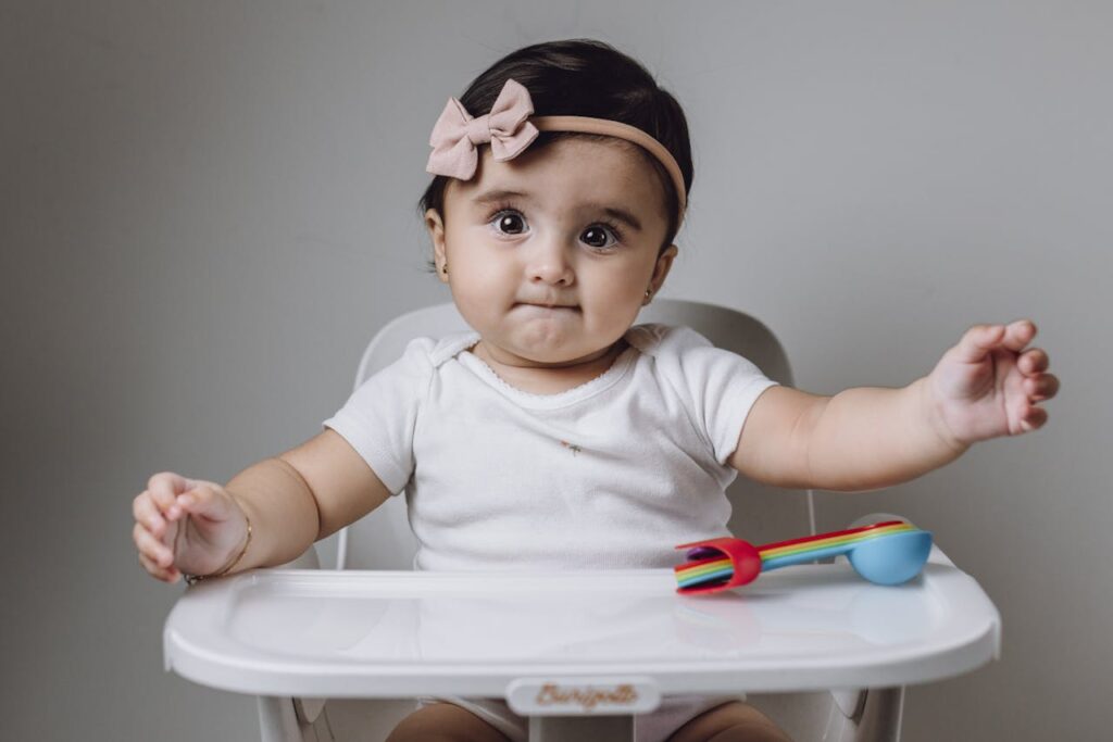 Cute baby girl in high chair with colorful toy utensils against gray background.